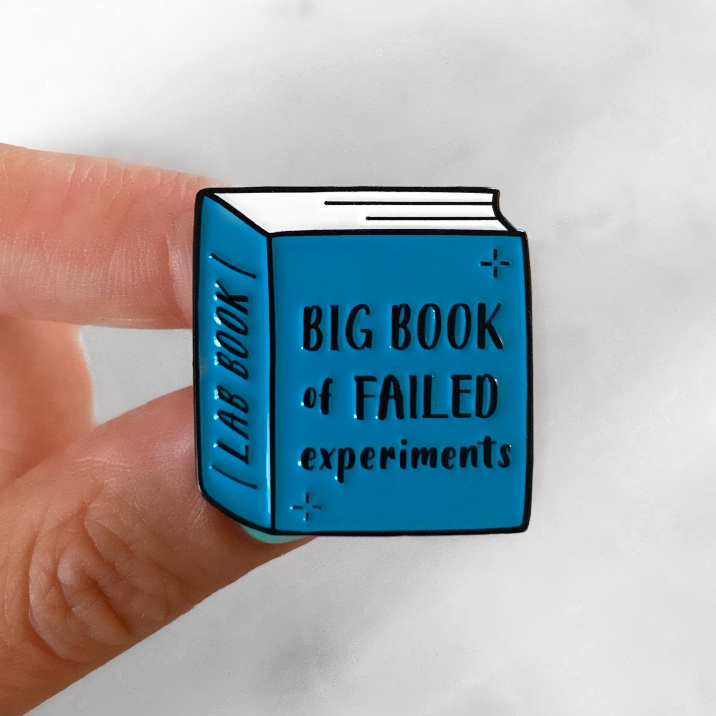 Blue enamel pin shaped like a book titled 'Big Book of Failed Experiments' held by a finger.