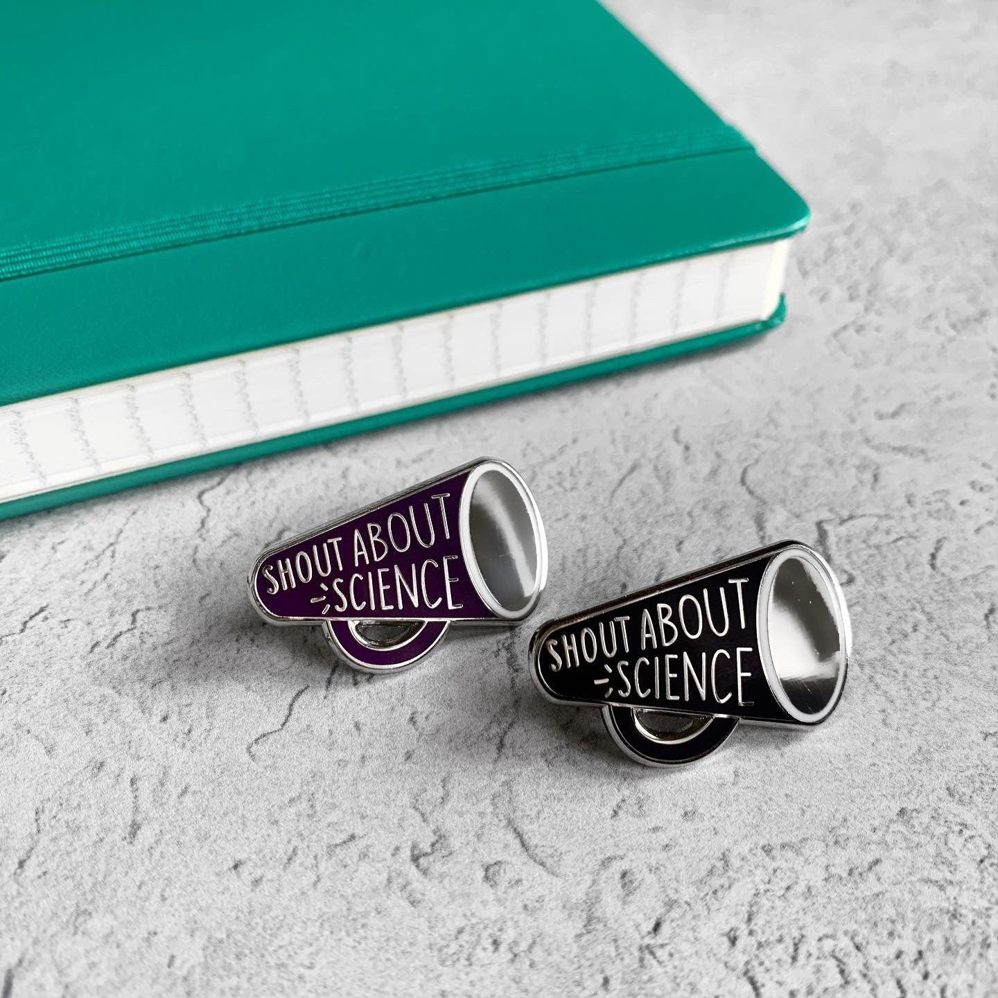 Two enamel pins with 'Shout About Science' text on a textured surface with a green notebook.