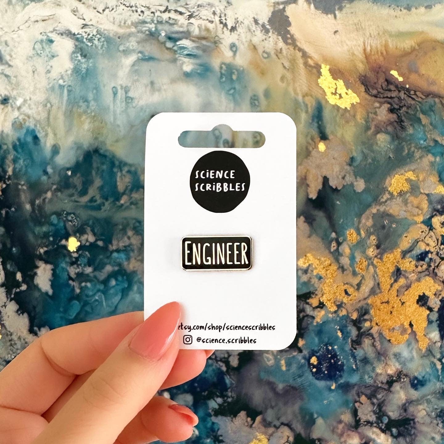 Hand holding a card with 'Science Scribbles' and 'Engineer' pins against a colorful abstract background.