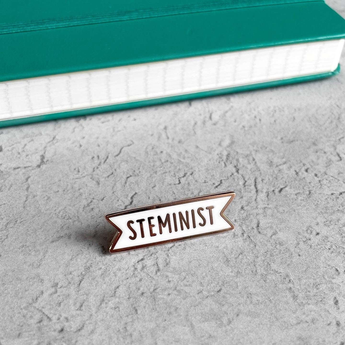 Enamel pin with 'STEMINIST' text on a gray surface with a teal book in the background