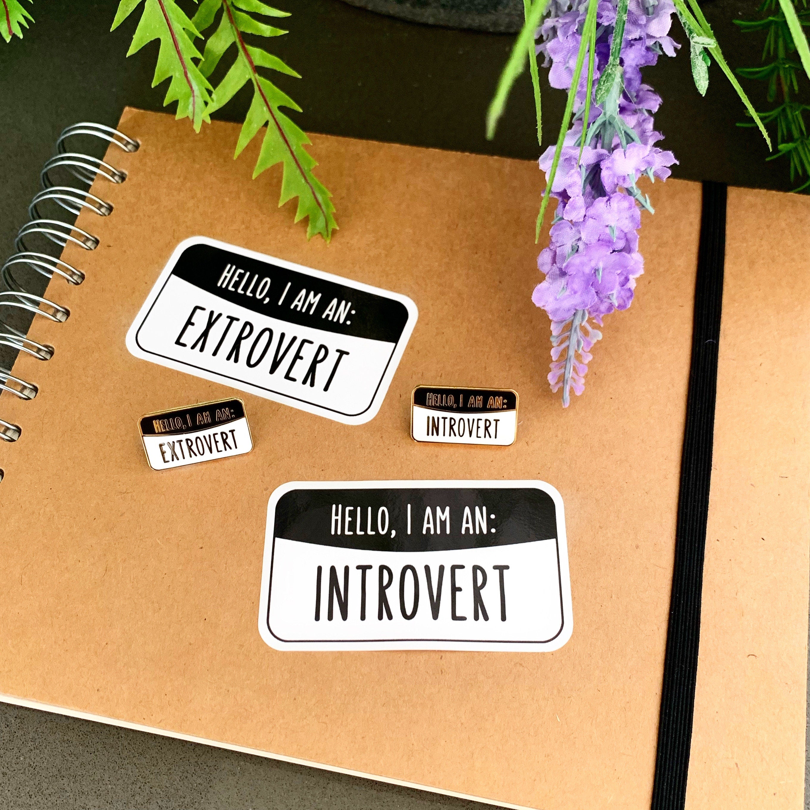 Notebook with 'Extrovert' and 'Introvert' stickers and enamel pins on a surface with lavender flowers.