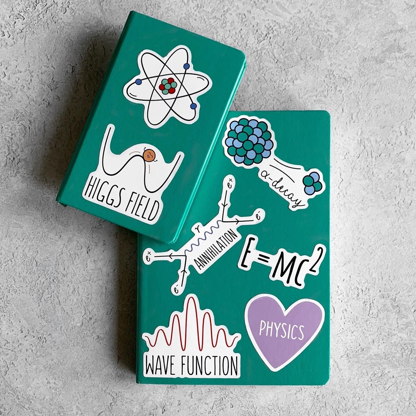 Two green notebooks with physics-themed stickers on a gray surface