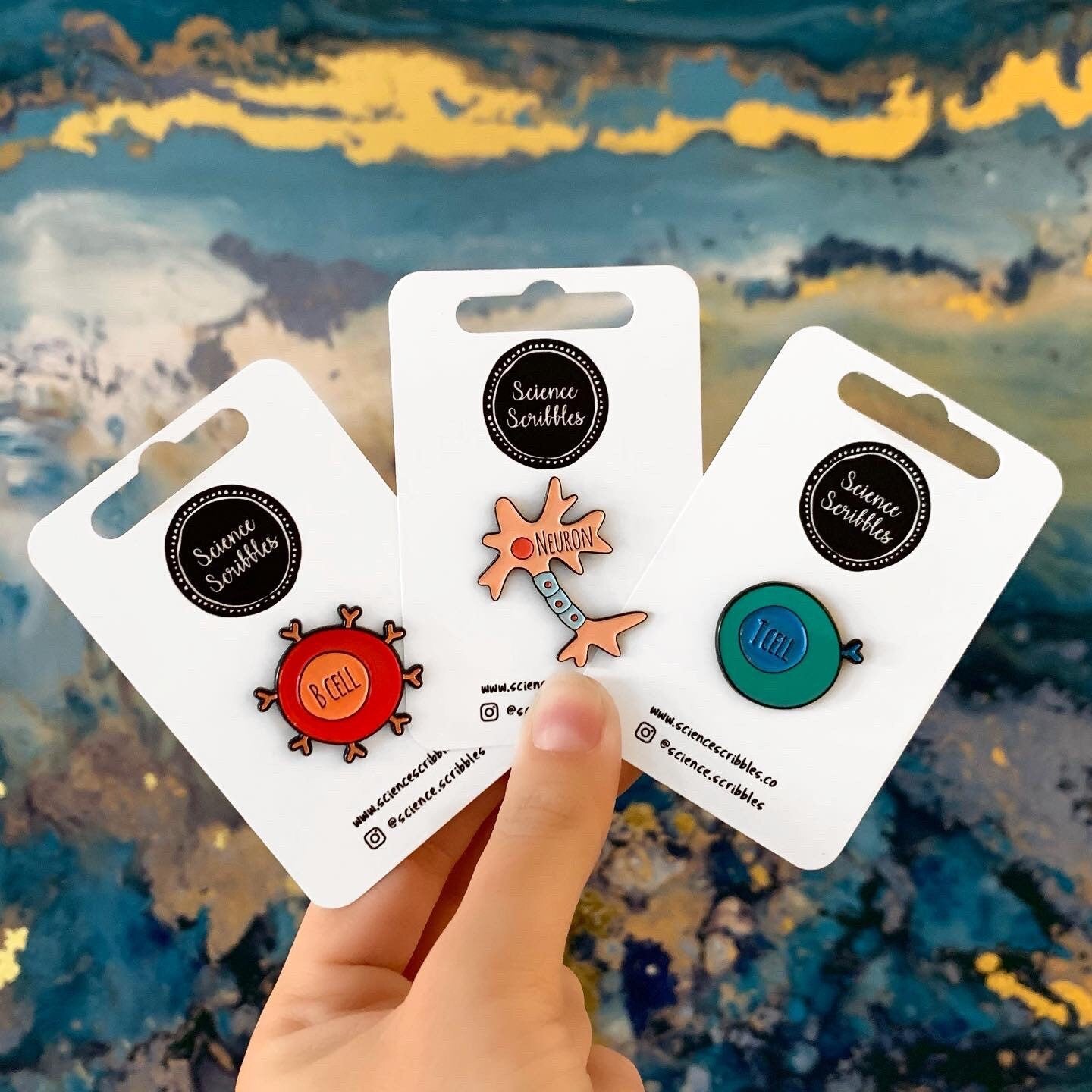 Three science-themed pins on cards held by a hand with a colorful background
