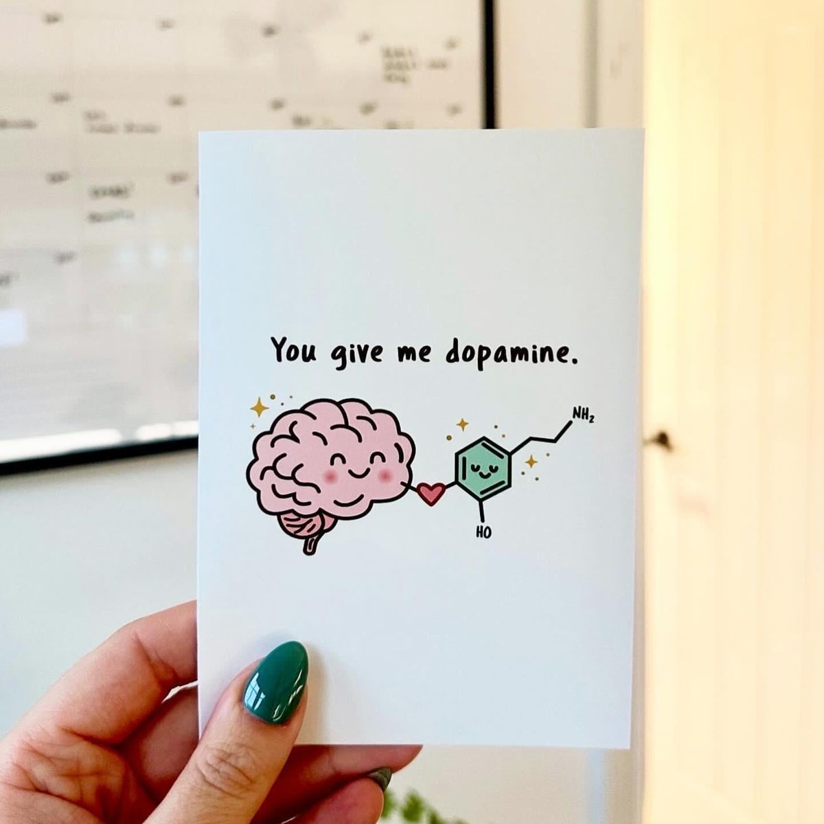A hand with green nail polish holds a white greeting card that reads 'You give me dopamine.' with a cartoon pink brain and a dopamine molecule illustration.