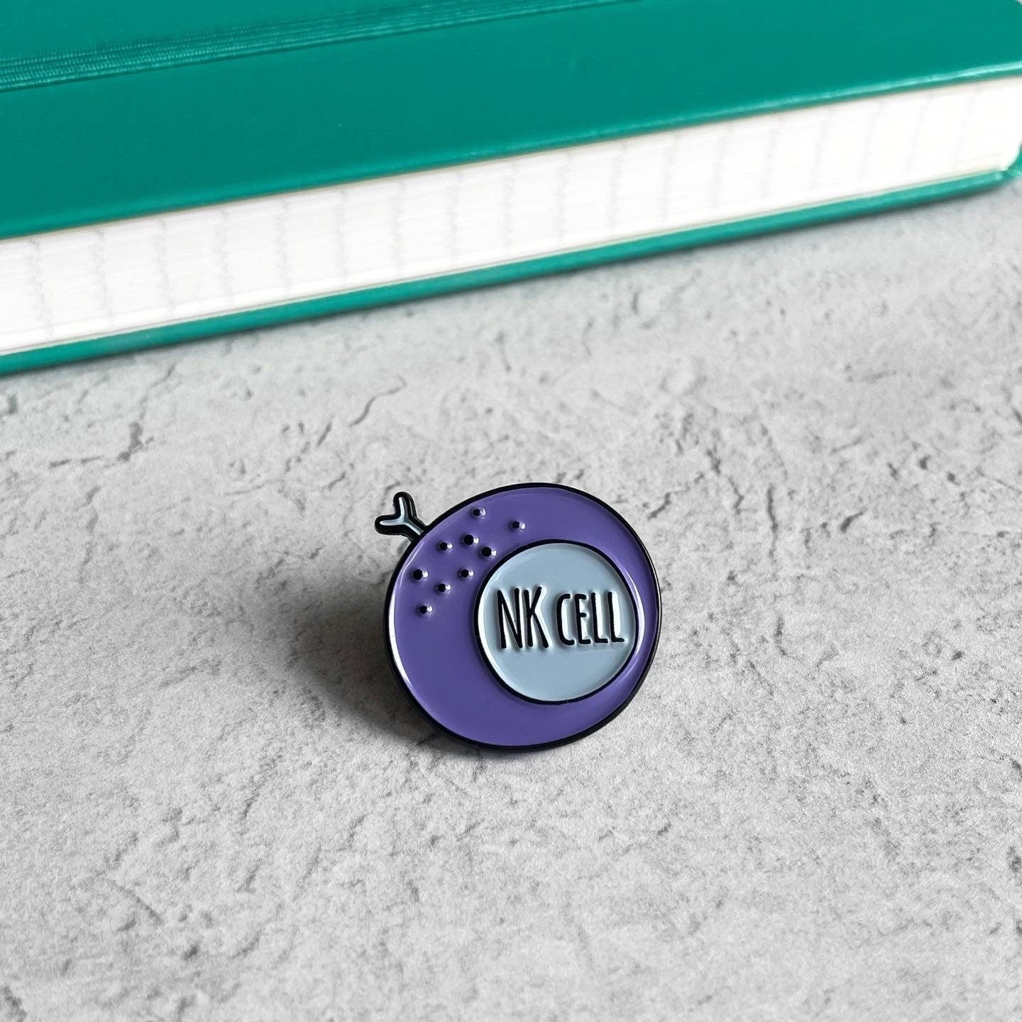 Purple and blue 'NK Cell' pin on a textured surface with a teal notebook in the background.
