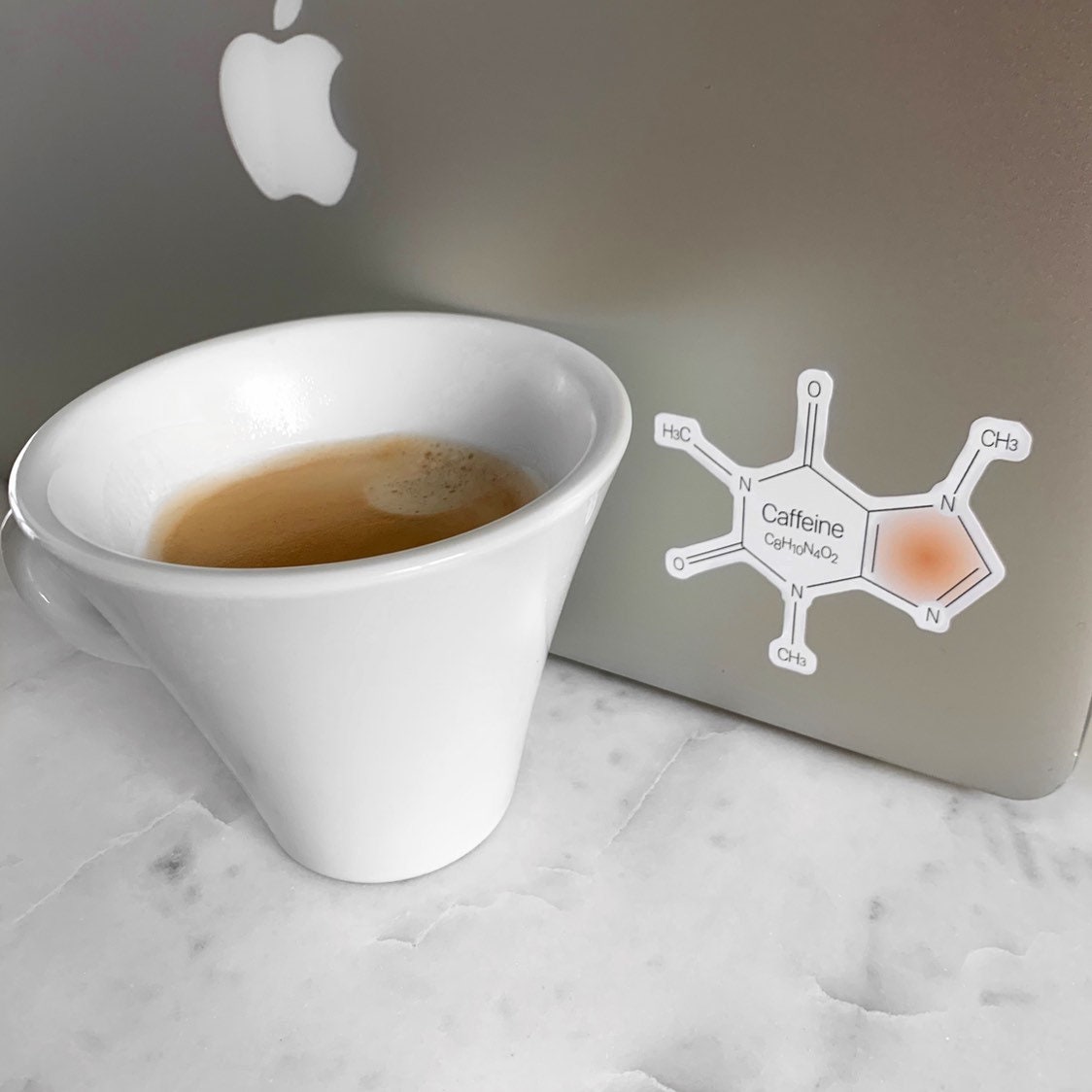 White mug with coffee next to a laptop with a caffeine molecule diagram sticker on a marble surface