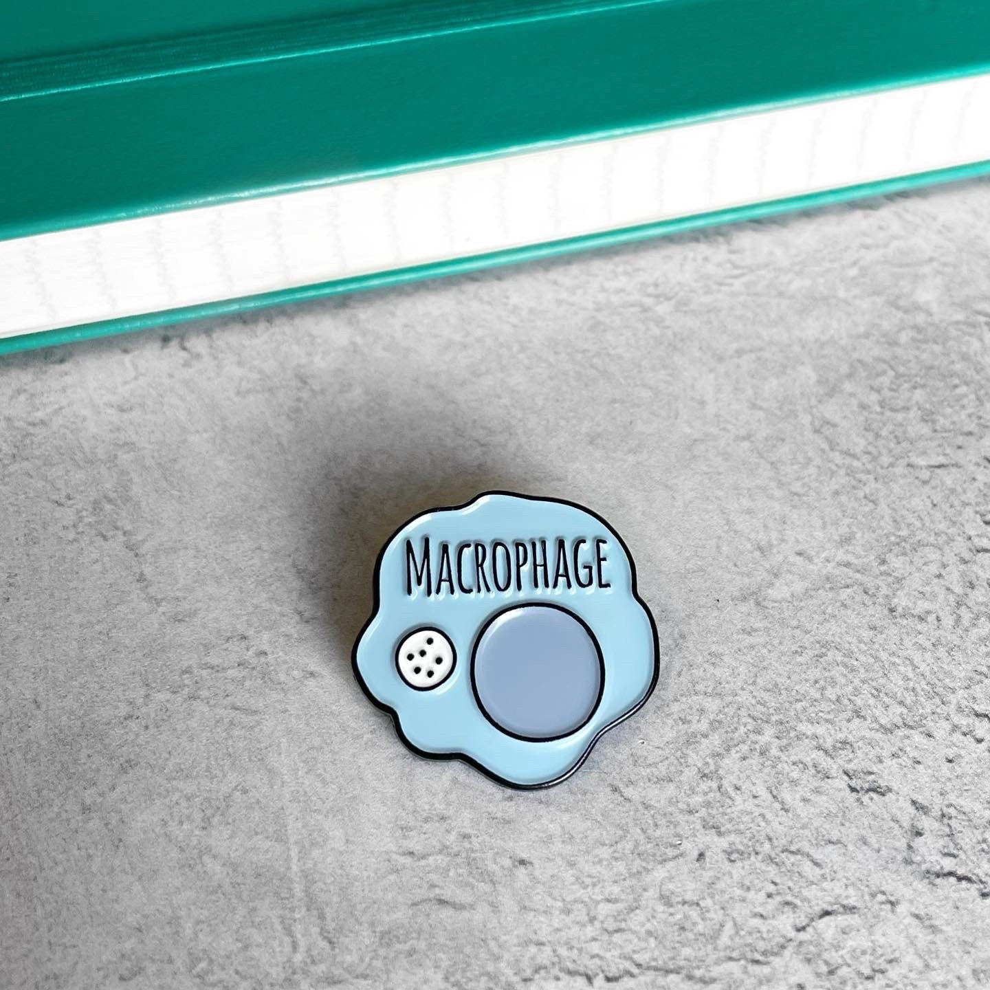 Enamel pin shaped like a macrophage on a textured surface with a blurred background