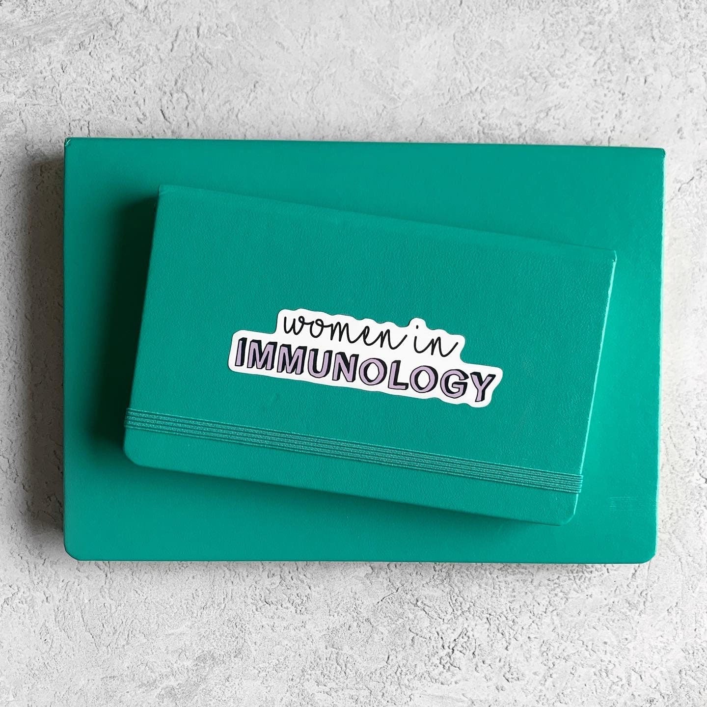 Teal notebook with 'women in IMMUNOLOGY' text on a light gray background