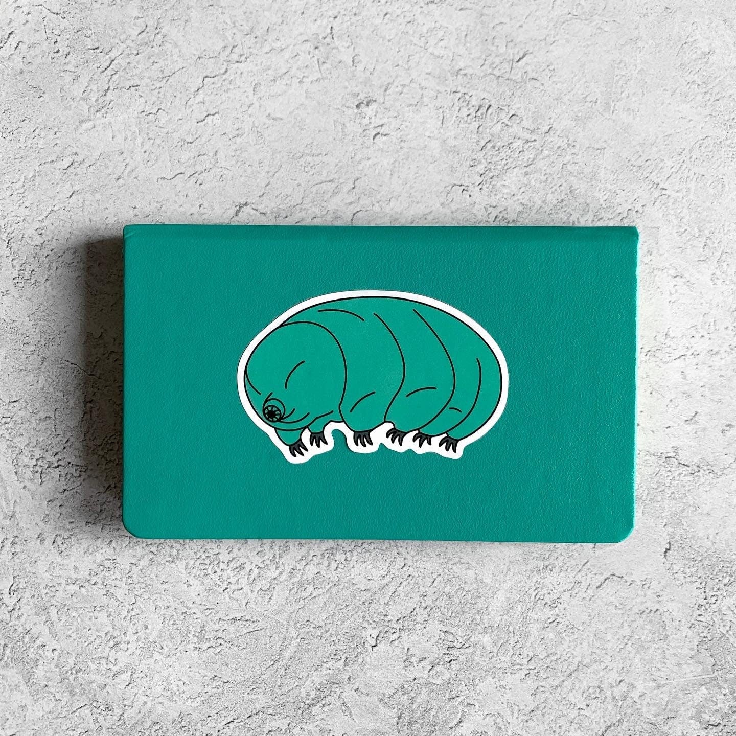Teal notebook with a white illustration of a tardigrade on a gray textured background