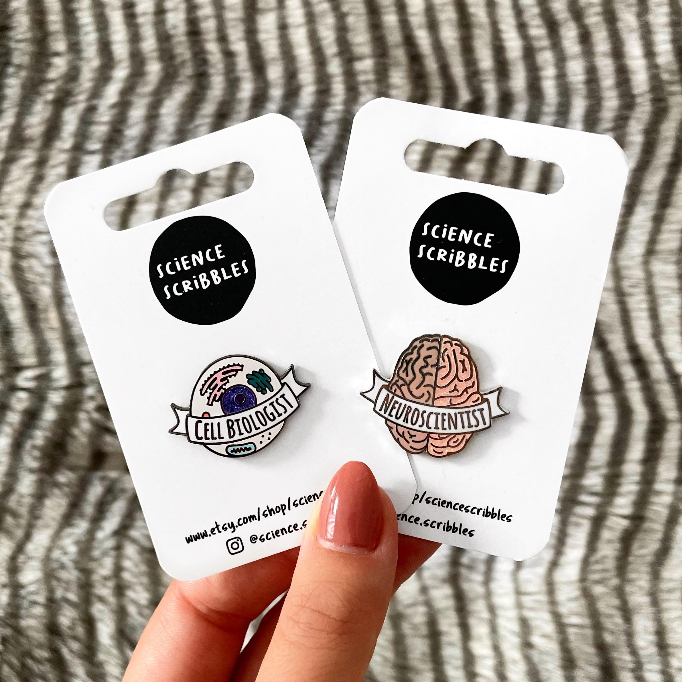 Two science-themed pins on 'Science Scribbles' cards held by a hand against a striped background.