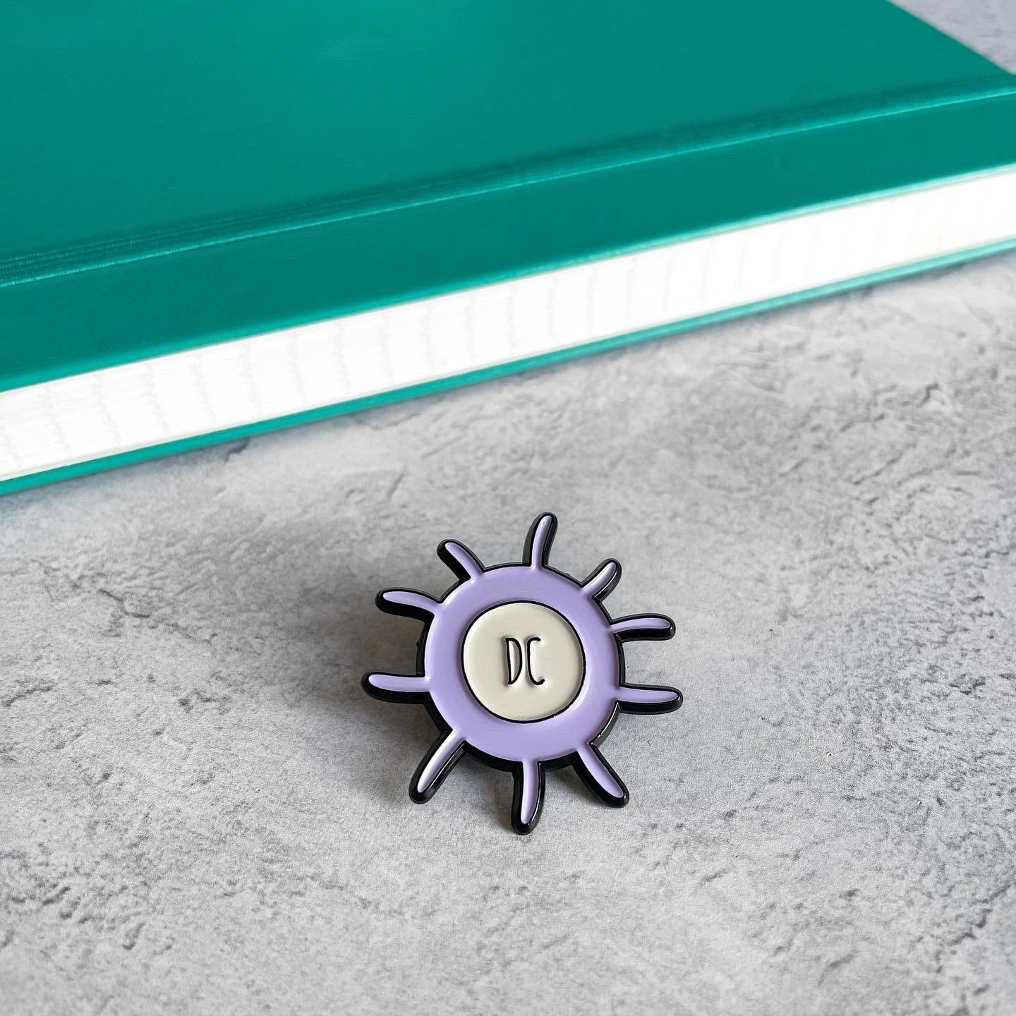 Dendritic cell purple and grey enamel pin badge on table with a teal notebook in the background