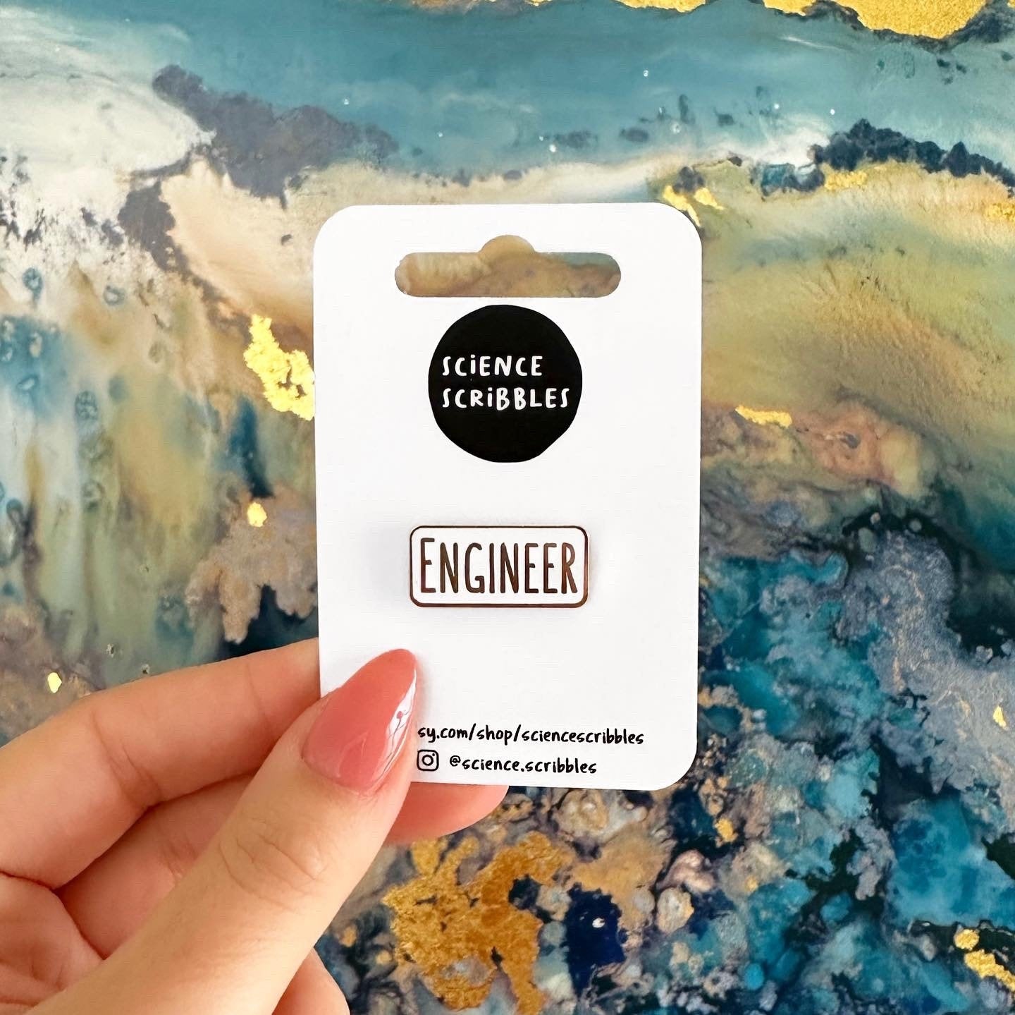 Hand holding a card with 'Science Scribbles' and 'Engineer' pins against a colorful abstract background.