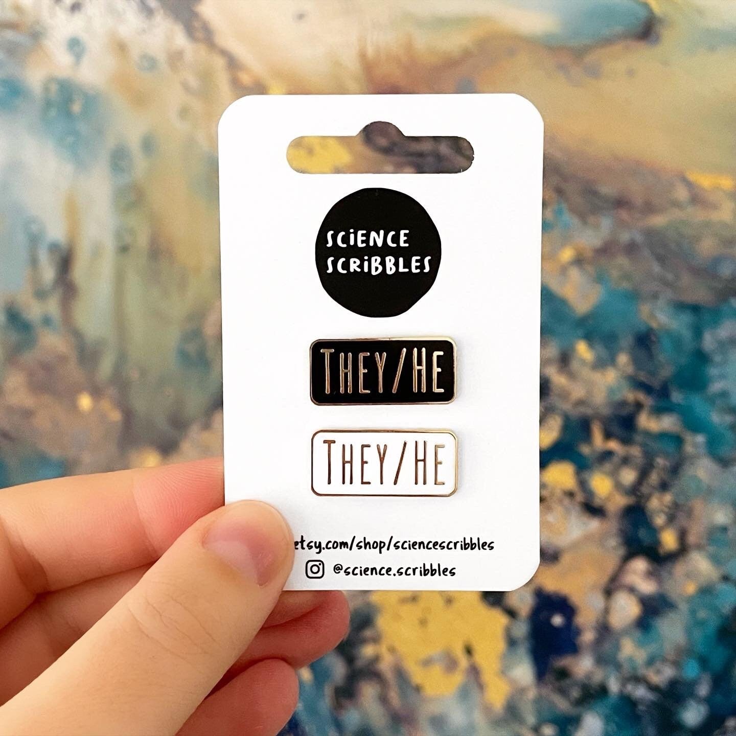 Hand holding a card with pronoun pins labeled 'SCIENCE SCRIBBLES' and 'THEY/HE'.