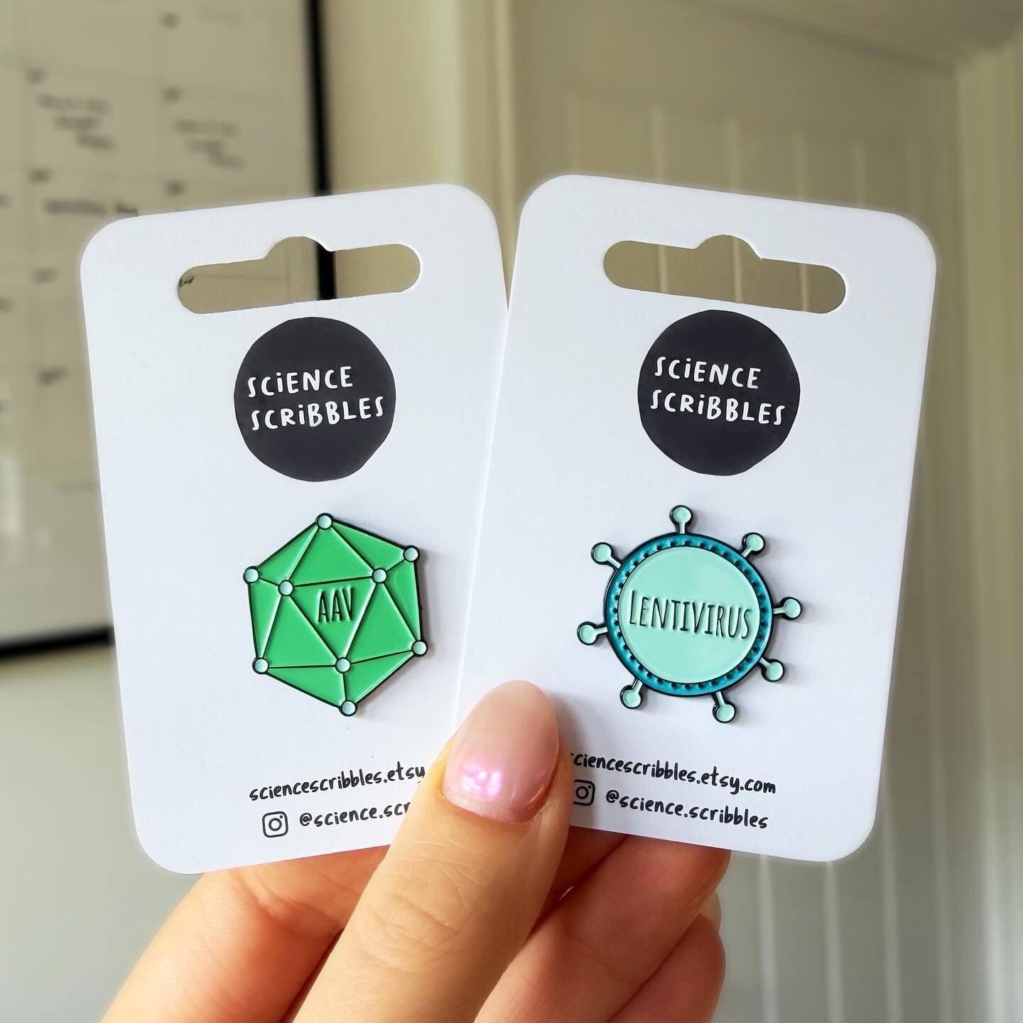 Two enamel pins on Science Scribbles cards held by a hand.