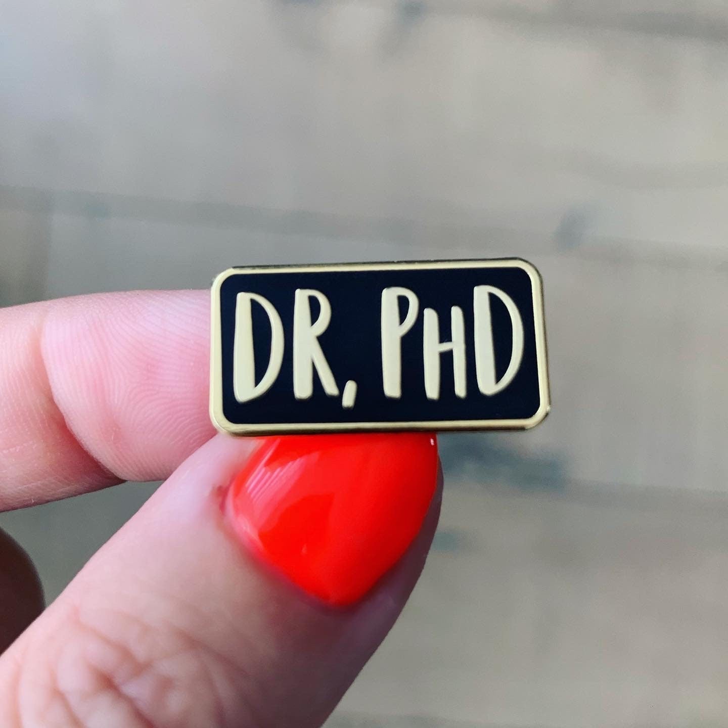 Pin with 'DR. PHD' text held by a hand with red nail polish.