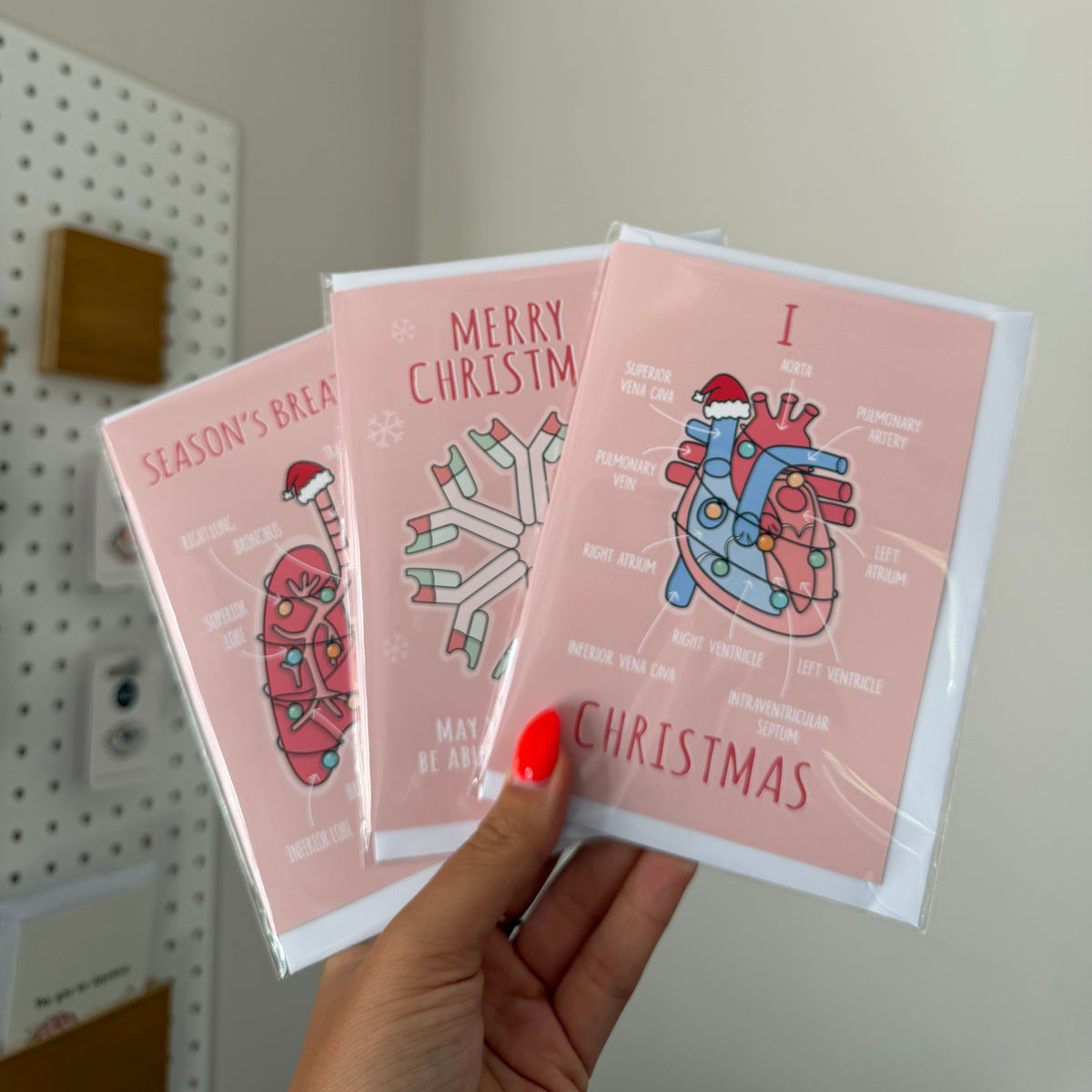 A hand with red nail polish holds three pink Christmas greeting cards with anatomical heart illustrations and holiday text, photographed indoors against a light wall.