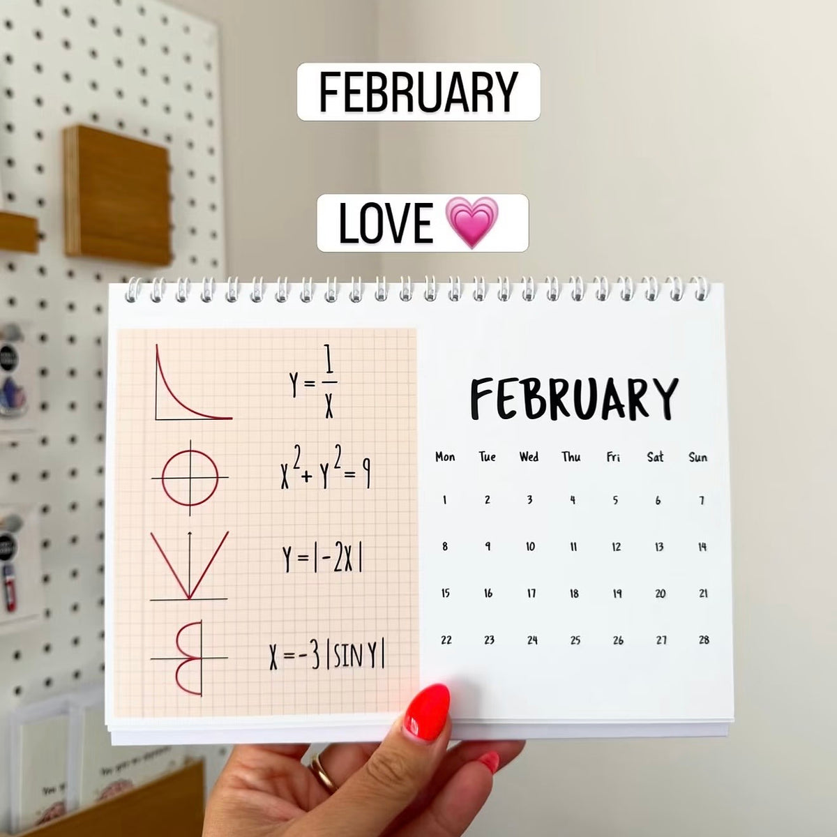 A hand with red nail polish holds a small spiral-bound February planner page featuring math equations on the left and a February calendar on the right. The background shows a pegboard wall with wooden shelves.
