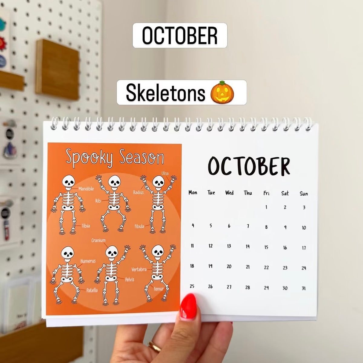 A hand with red nail polish holds a spiral-bound October desk calendar featuring a 'Spooky Season' page with cartoon skeletons and a standard October calendar grid. Text labels 'OCTOBER' and 'Skeletons' appear above.