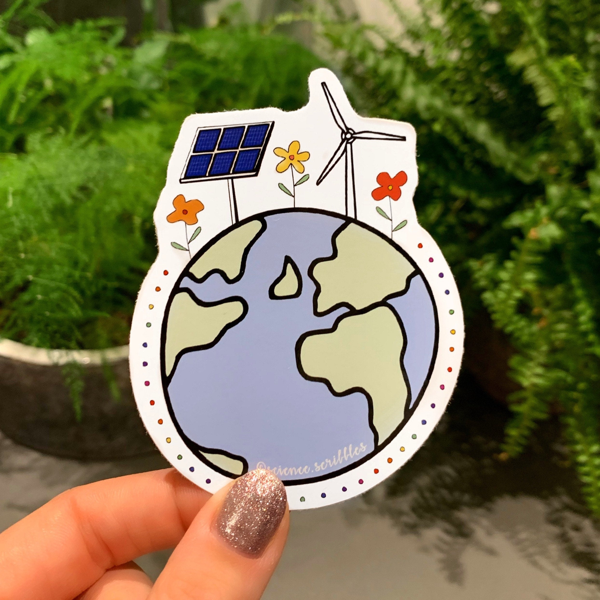 Sticker of a globe with solar panel, wind turbine, and flowers held by a hand against a green plant background.