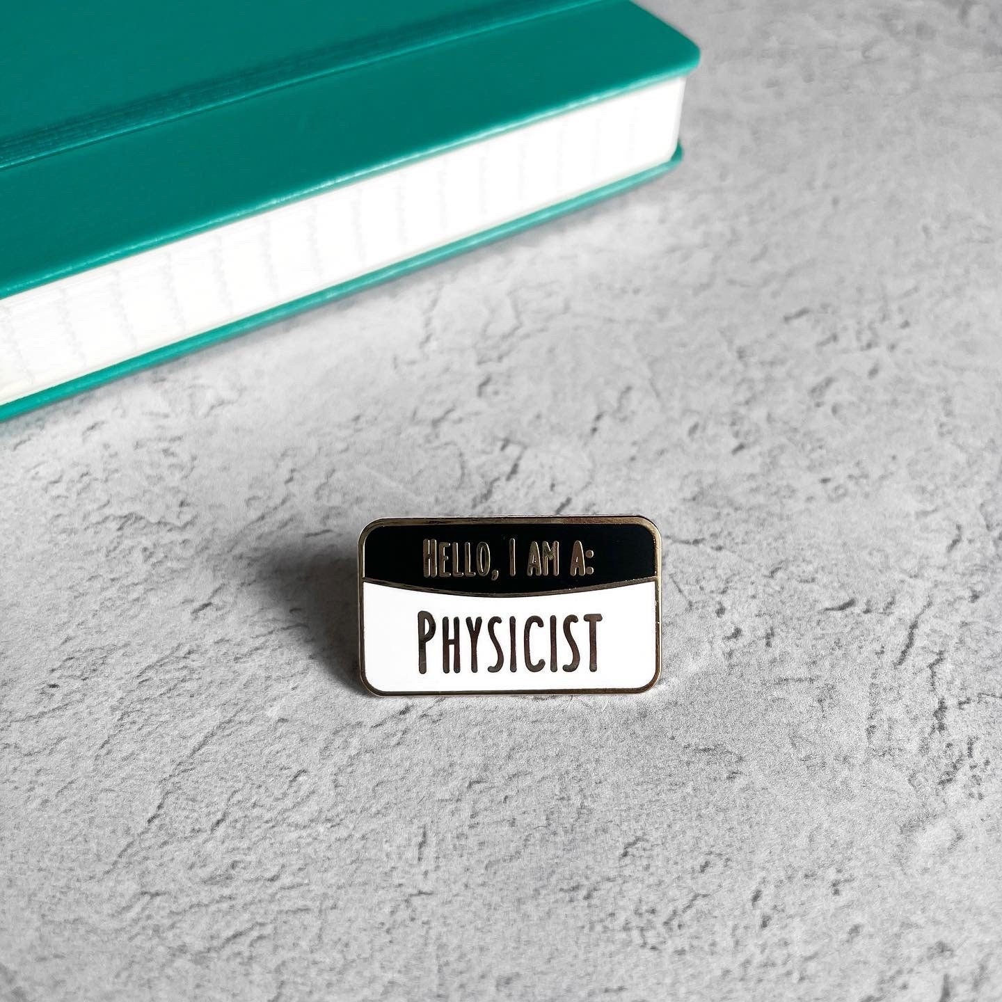 Pin with 'Hello, I am a Physicist' text on a textured surface with a book in the background