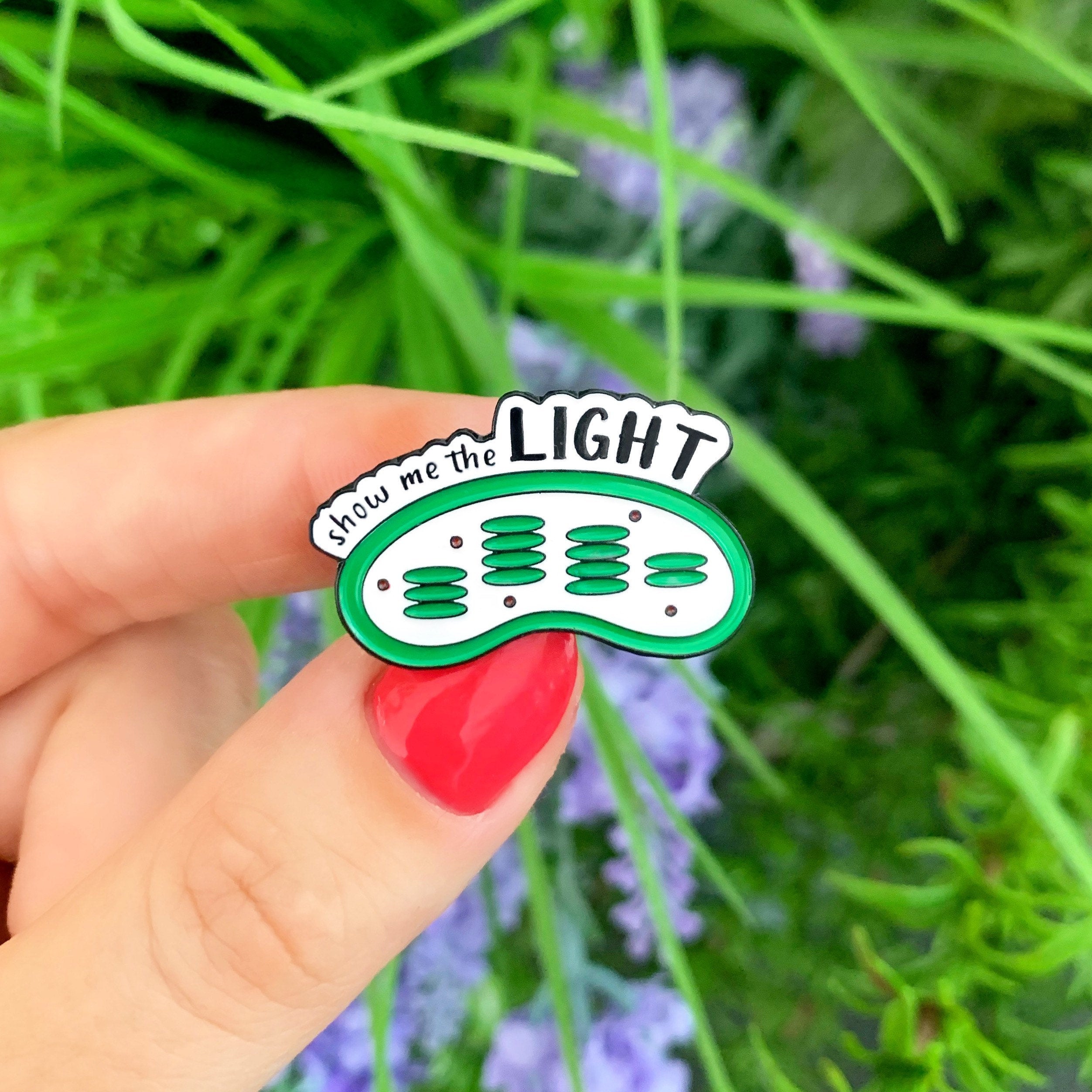 Hand holding a green and white enamel pin with 'Show me the Light' text against a blurred green and purple plant background.