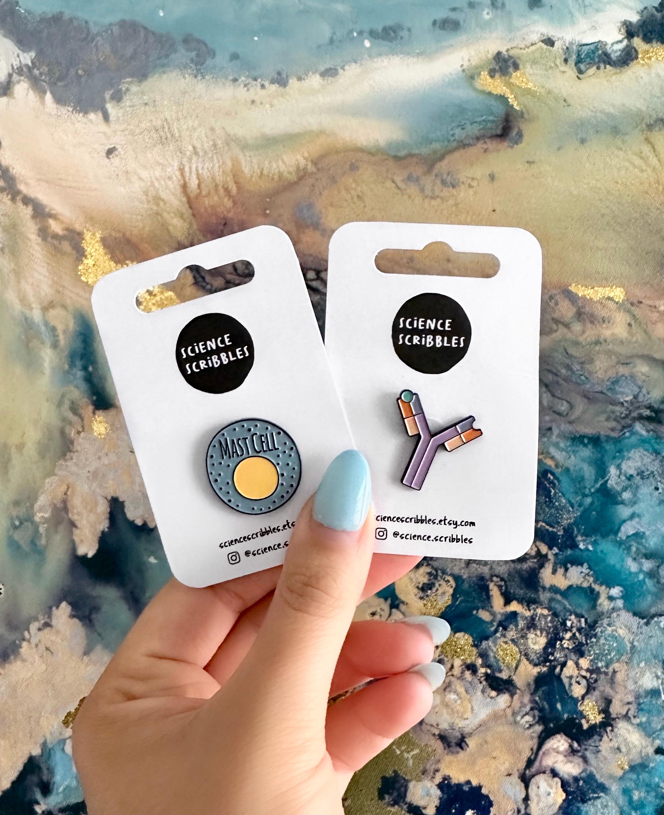 Two packages of enamelpins held by a hand against a textured background