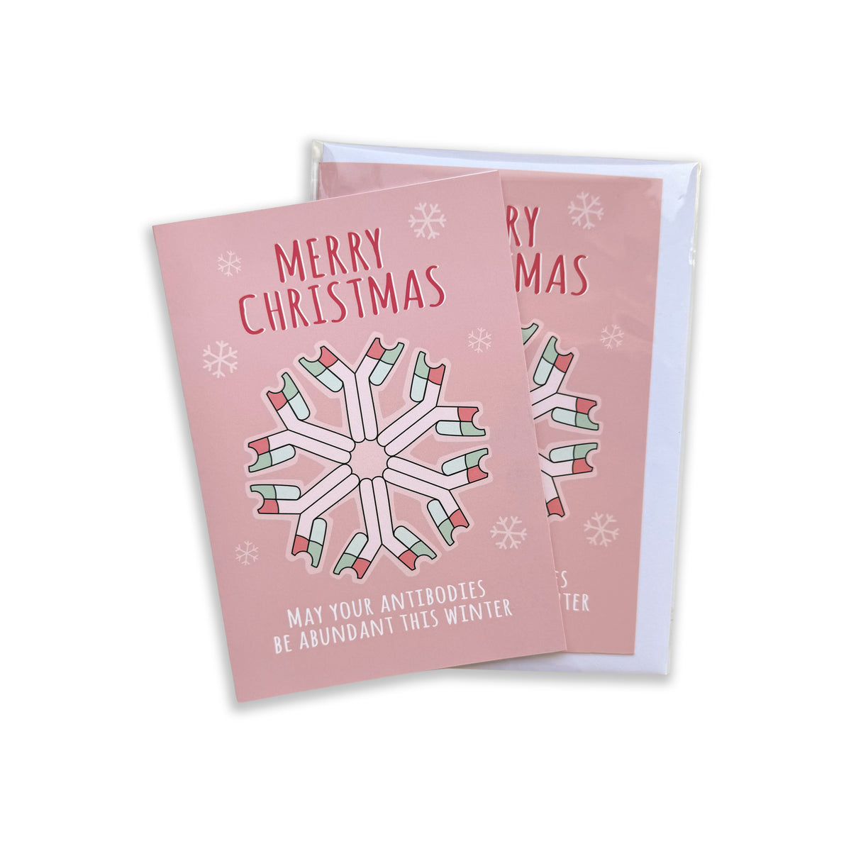 A pink Christmas greeting card with a snowflake made of antibiotics and the text 'MERRY CHRISTMAS' and 'MAY YOUR ANTIBIOTIDES BE ABUNDANT THIS WINTER', shown with a white envelope on a white background.