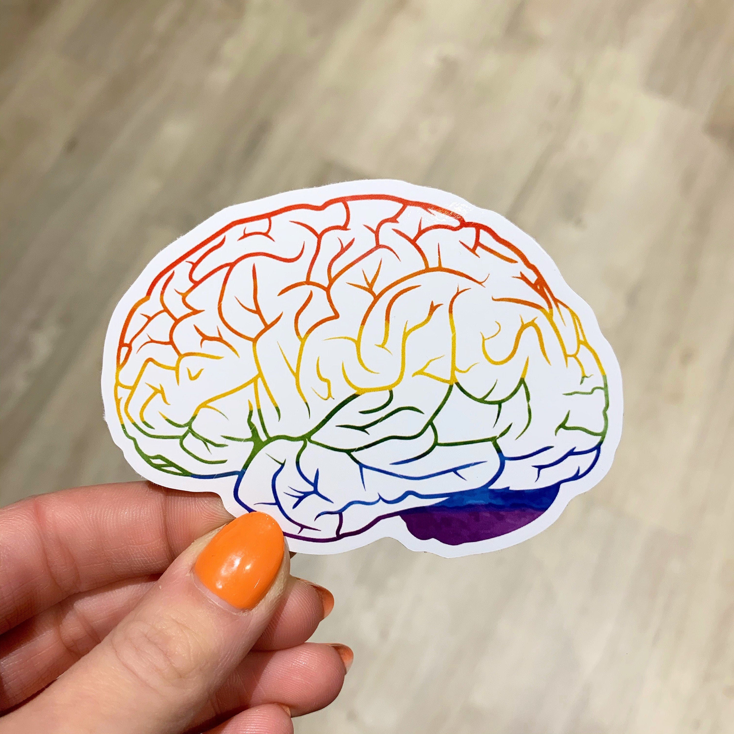Colorful brain sticker held by a hand with orange nail polish on a wooden surface