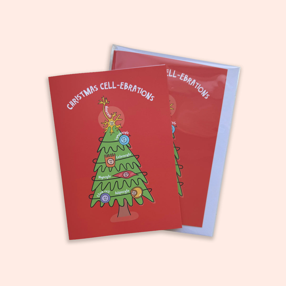 A red Christmas greeting card with a cartoon tree made from cell parts, topped with a giraffe and decorated with colorful round ornaments, shown with a light blue envelope on a pale peach background.