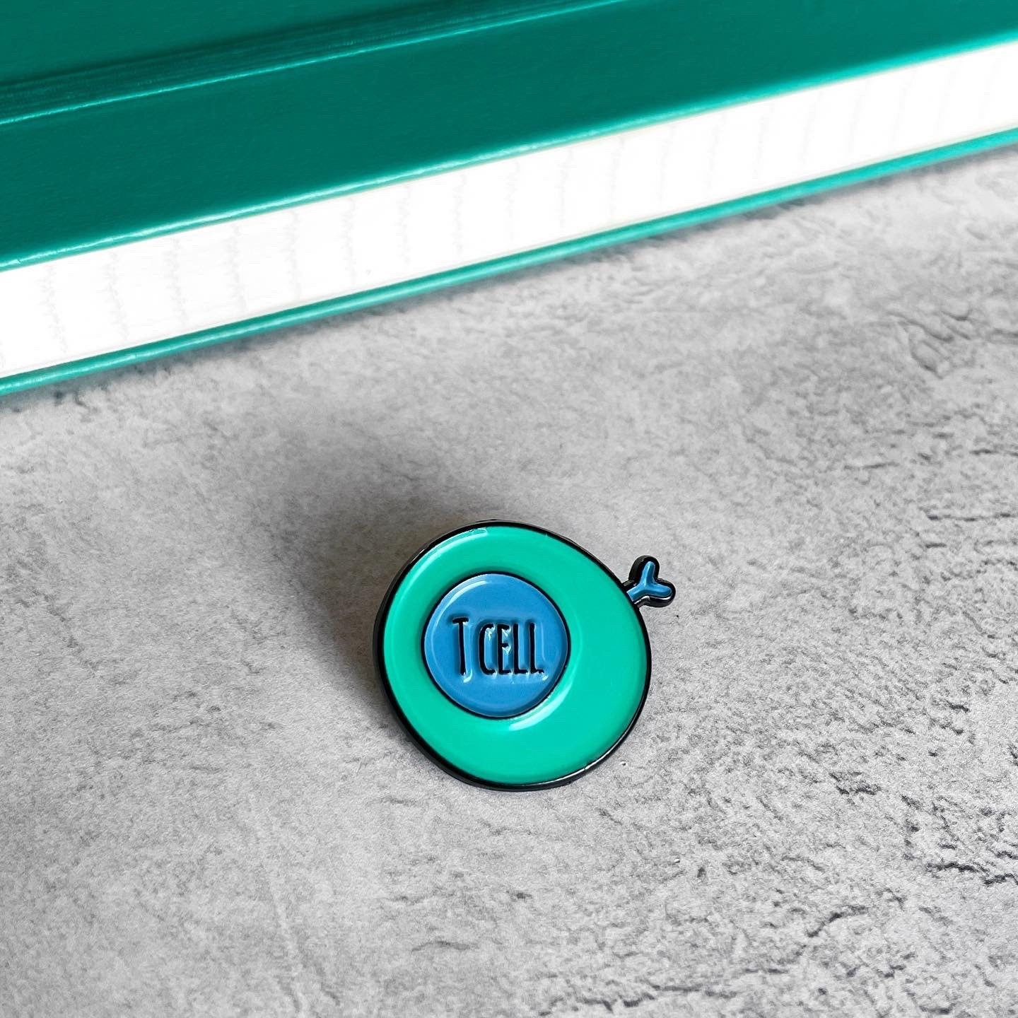 Round blue and green enamel pin with 'T cell' written inside.