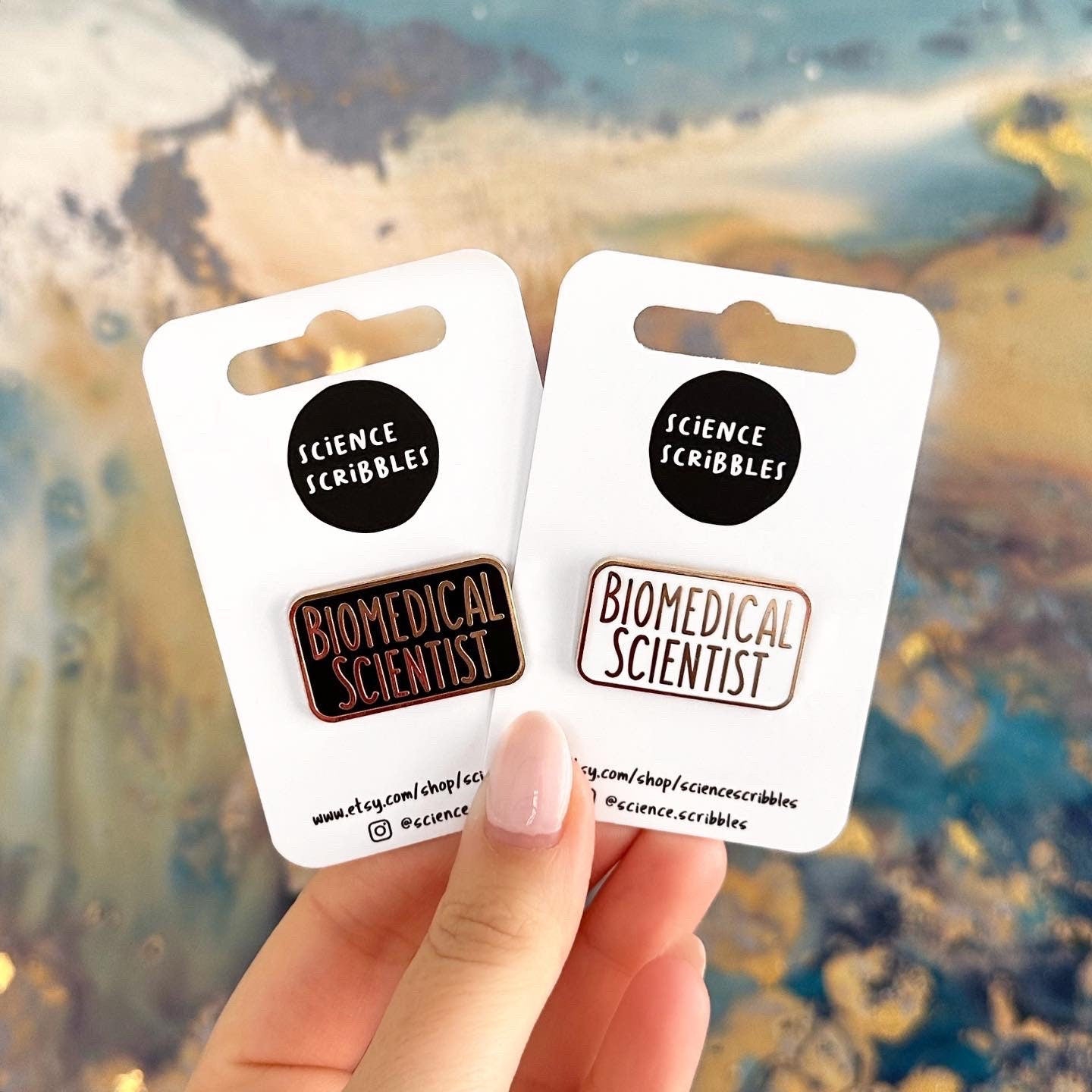 Two enamel pins with the text 'Biomedical Scientist' on it. One is white and gold and the other is black and gold.