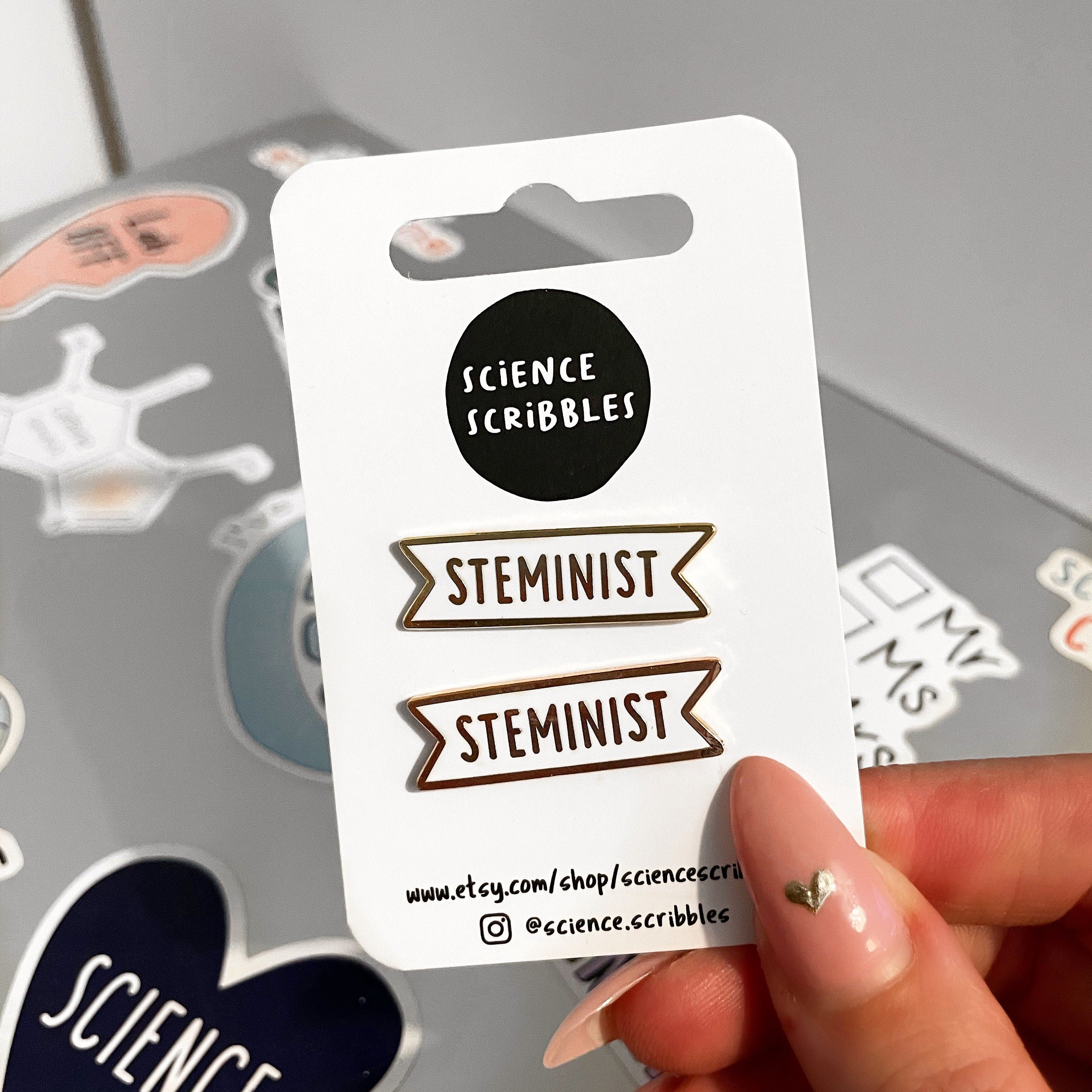 Packaging of 'STEMINIST' enamel pins from Science Scribbles held by a hand.
