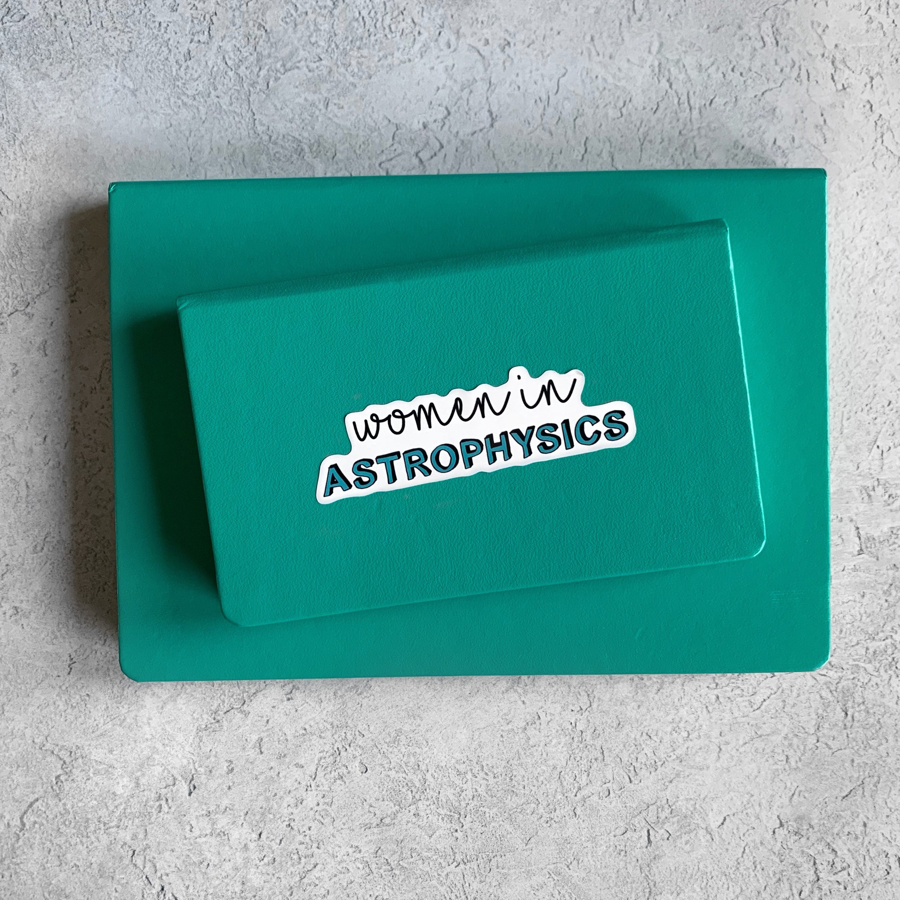 A teal notebook with 'women in astrophysics' text on a gray surface