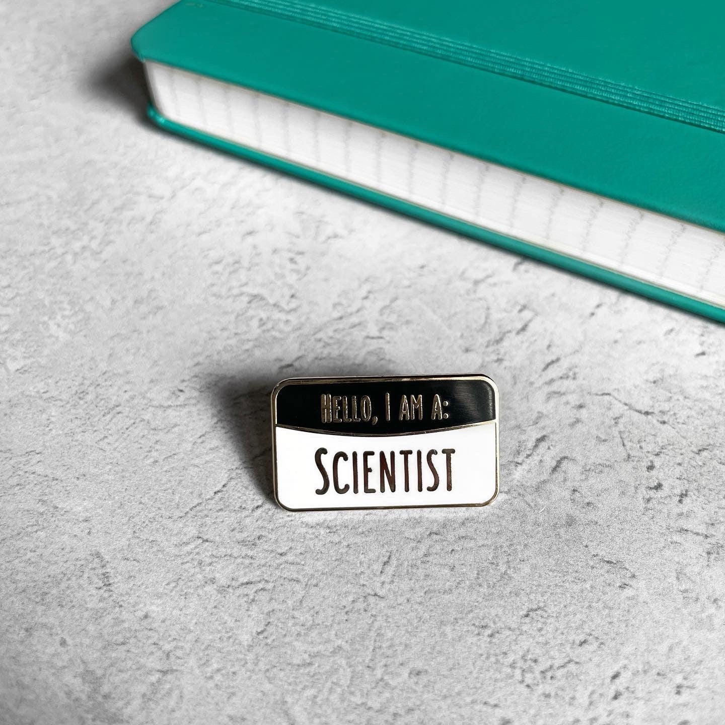 Pin with 'Hello, I am a Scientist' text on a gray surface with a teal notebook.