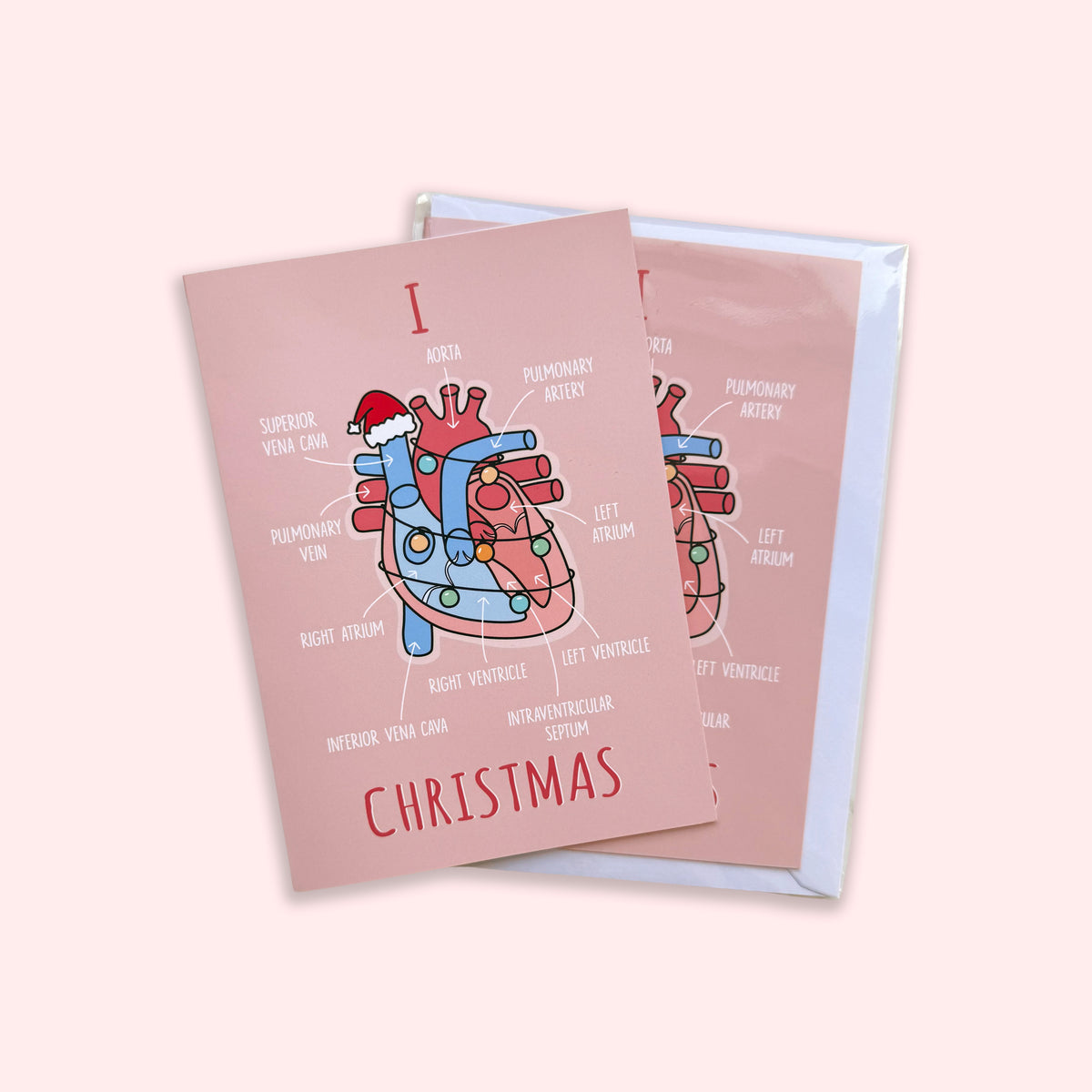Two pink Christmas greeting cards with an anatomical heart illustration and the word 'CHRISTMAS' printed on them, shown on a pale pink background.