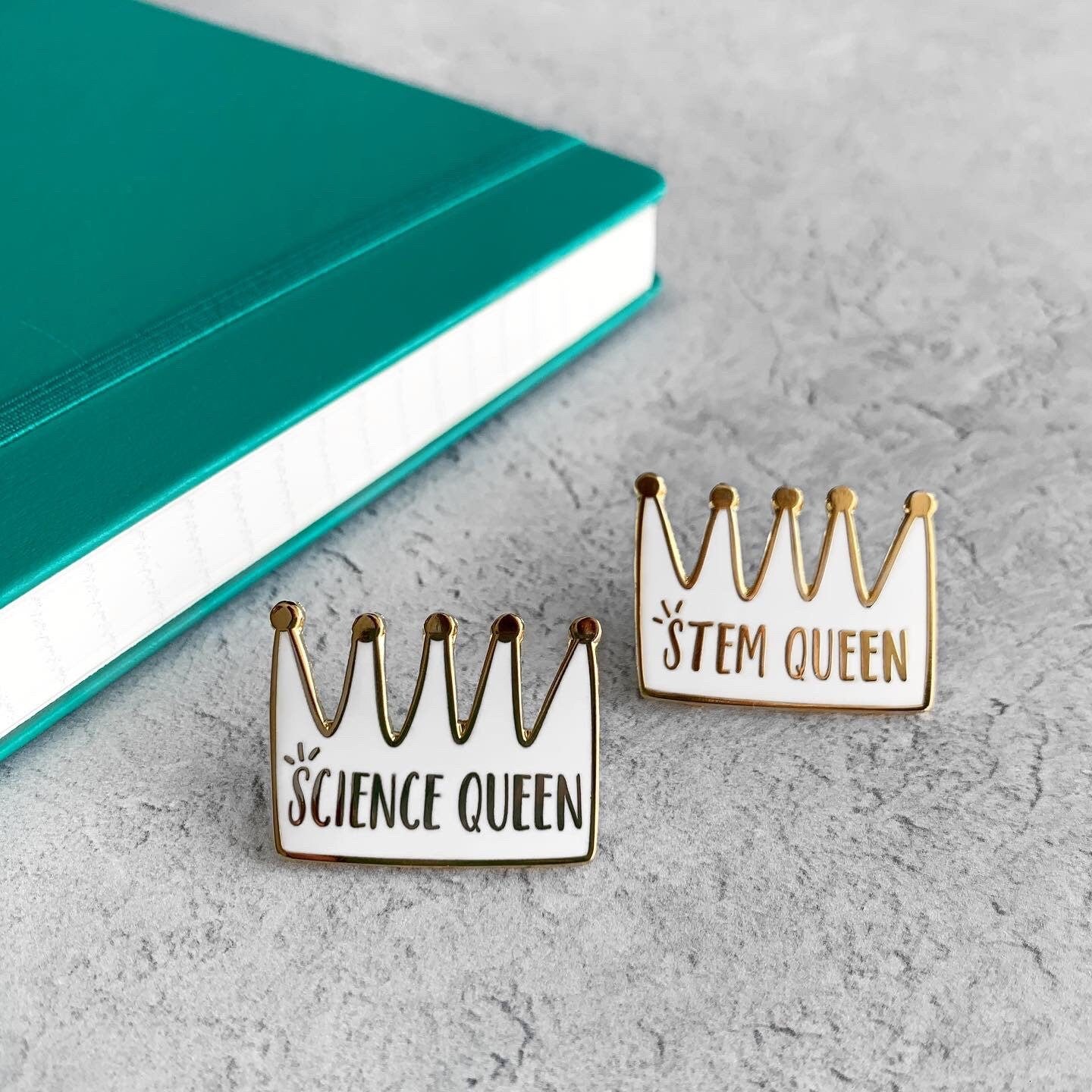 Crown-shaped pins with 'STEM Queen' and 'Science Queen' text on a gray surface with a teal notebook.