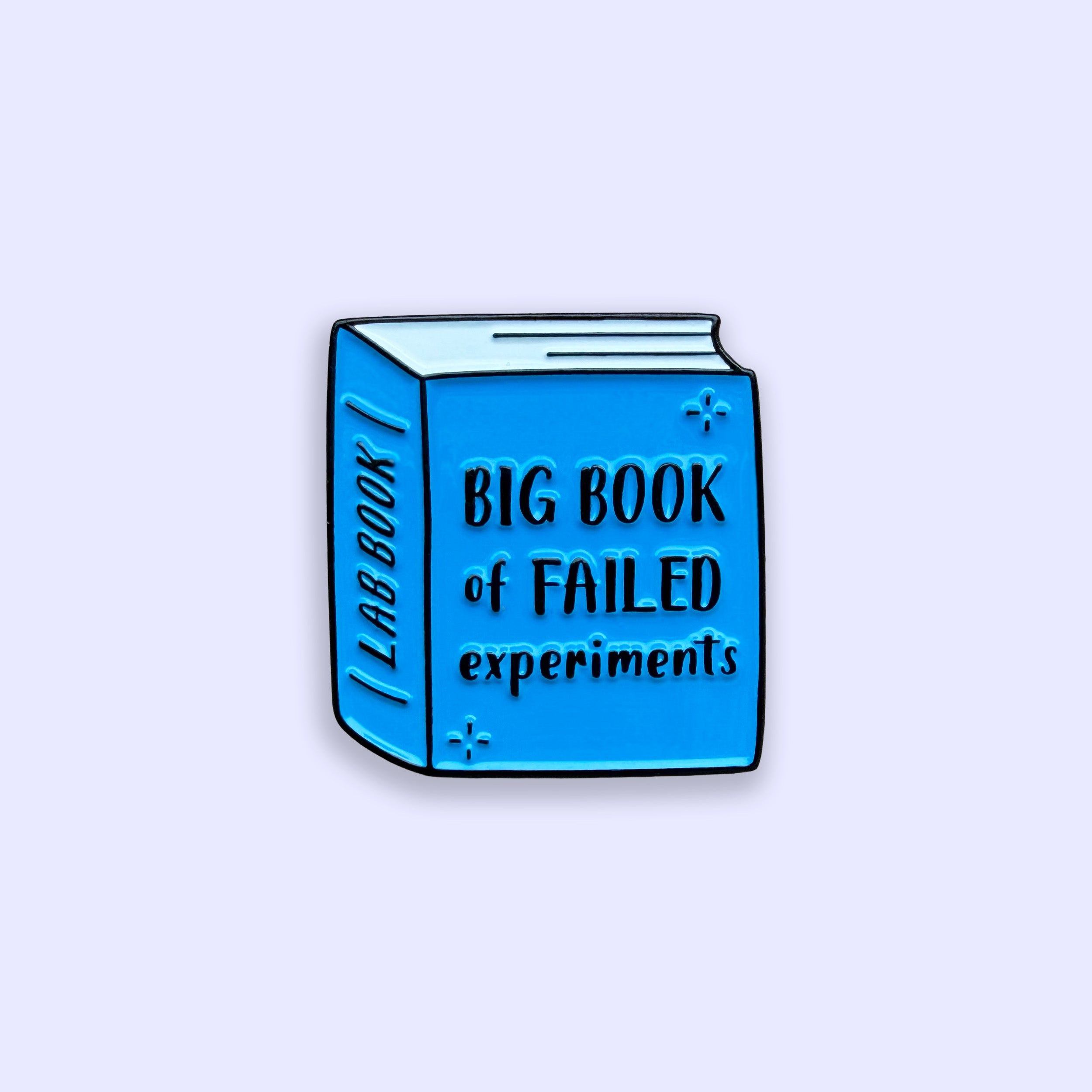 Blue enamel pin shaped like a book titled 'Big Book of Failed Experiments' on a light purple background