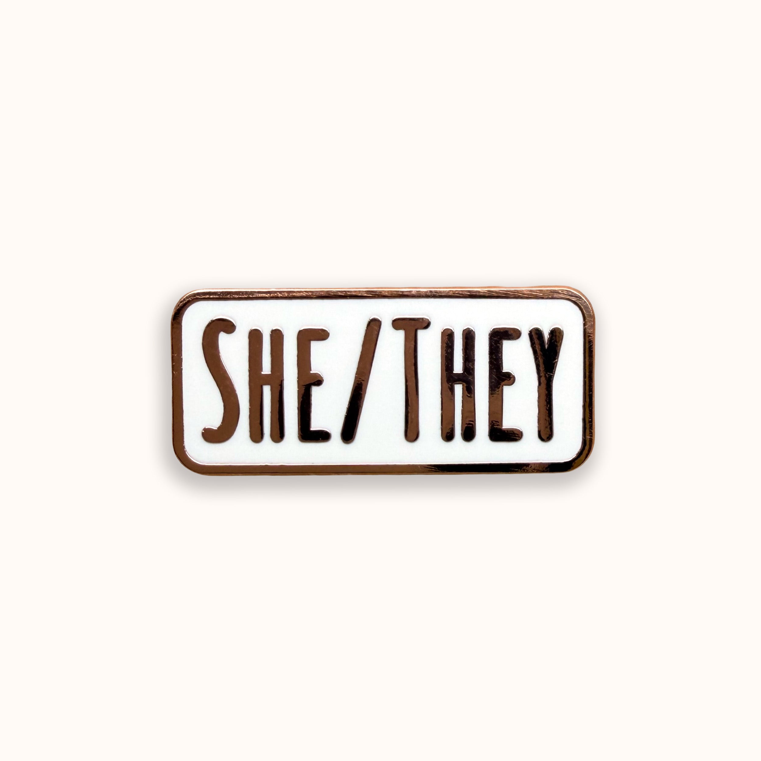 She/They pronoun white and rode gold enamel pin badge on a pink background
