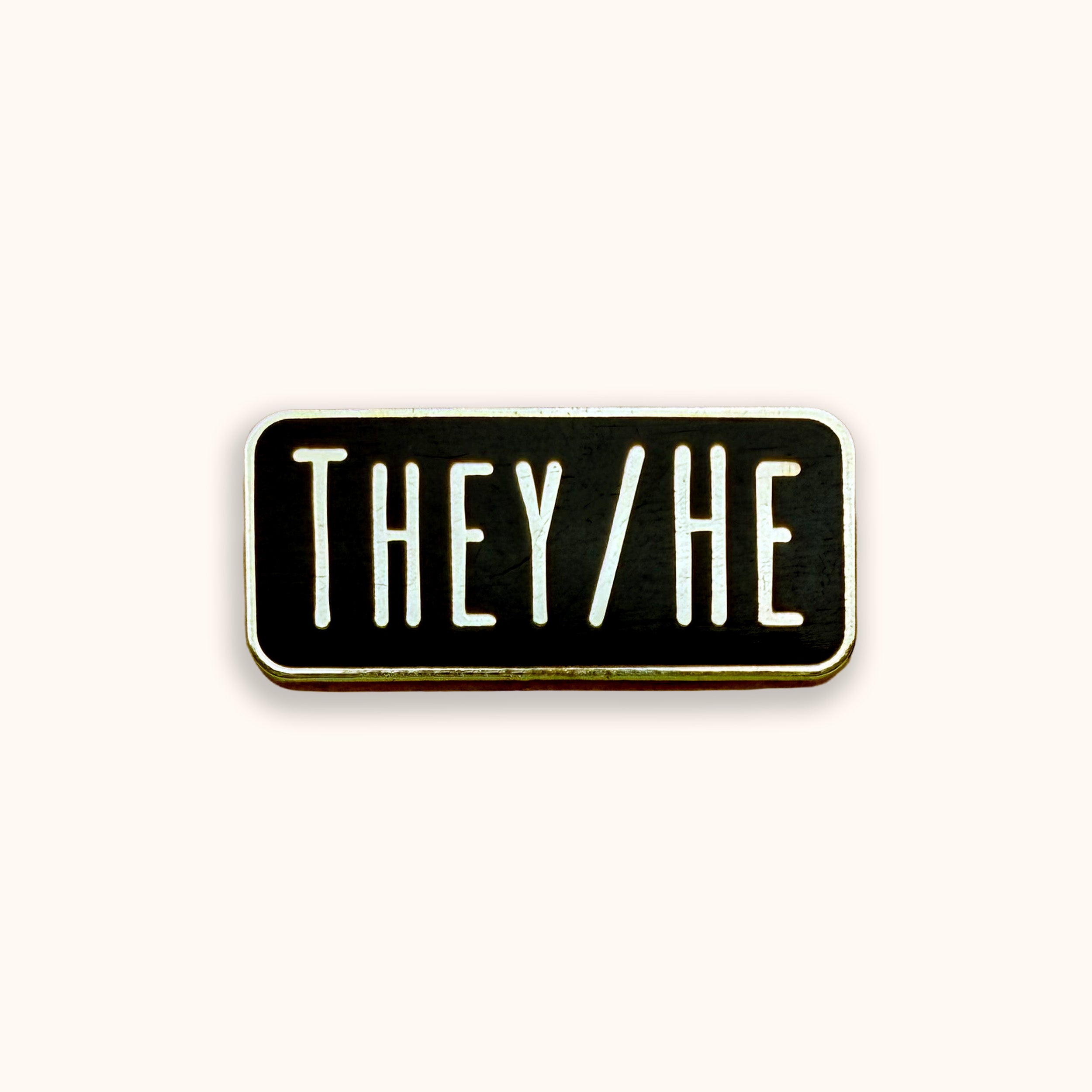 A rectangular enamel pin with a black face, white text reading 'THEY/HE', and a gold-toned border, centered on a light cream background.